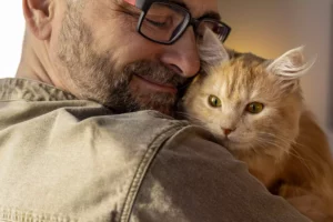 Auf dem Foto ist ein Patrick mit Brille zu sehen, der liebevoll seine orangefarbene Hauskatze Maria im Arm hält. Patrick hat die Augen geschlossen und lächelt sanft, was eine ruhige und innige Verbindung zwischen ihm und Mariavvermittelt. Die Katze blickt aufmerksam und entspannt nach vorne, während sie sich dicht an Patrick schmiegt. Beide wirken zufrieden und geborgen. Das Licht ist warm und weich, was die emotionale, vertraute Stimmung des Moments unterstreicht.
